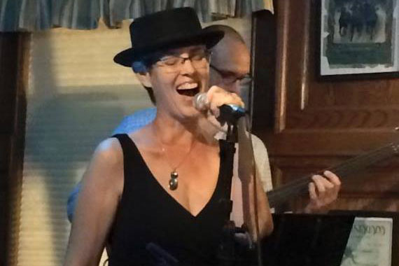 Vocalist Kim Scudera of Batida Diferente dressed in a black dress with a black hat, sings on the Henry's stage.